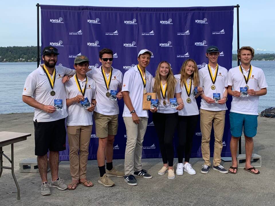 Eight young people wearing white shirts and medals stand in a row smiling in front of a blue NWISA banner by the water, holding plaques and awards.