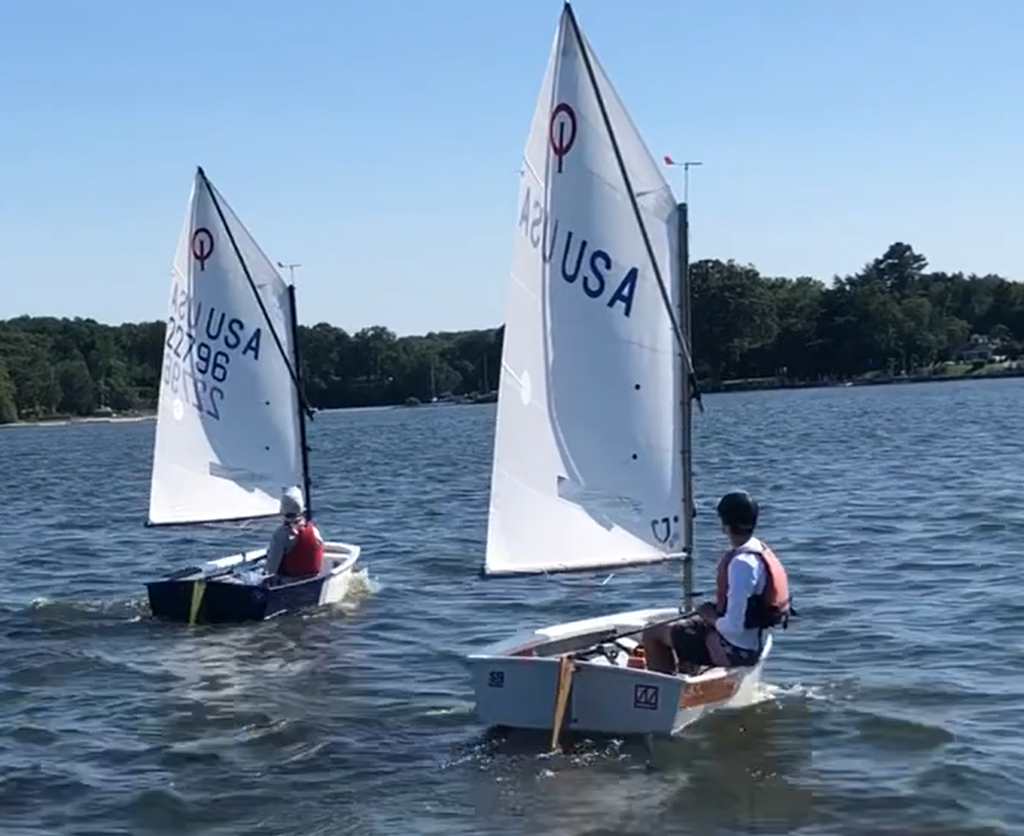 Two young sailors in small boats with white sails marked "USA" sail on a lake under a clear blue sky, with trees visible along the shoreline in the background.