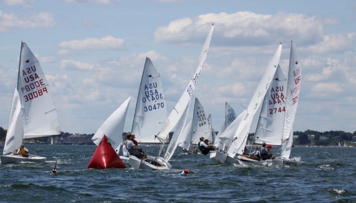 Several small sailboats with visible sail numbers race around a red buoy on a sunny day, with the shoreline and cloudy sky in the background.