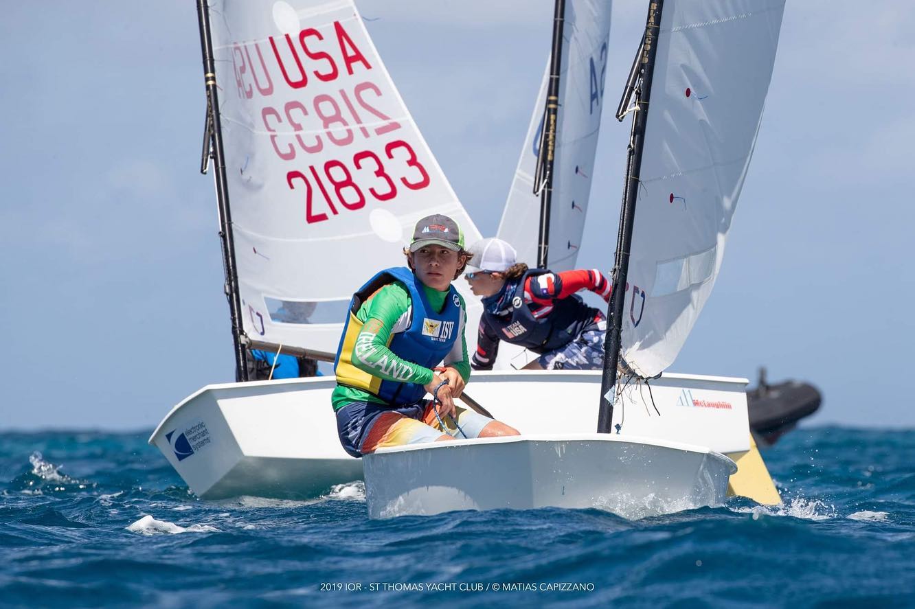 Two young sailors compete in small dinghy sailboats on blue water, with one wearing a green and yellow vest and the other in a dark top. The sails display numbers and "USA." The sky is clear and sunny.
