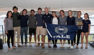 A group of twelve people smile and pose together indoors by the water. Several are holding a large blue Yale flag, and one person holds a wooden plaque. They appear to be celebrating an achievement.