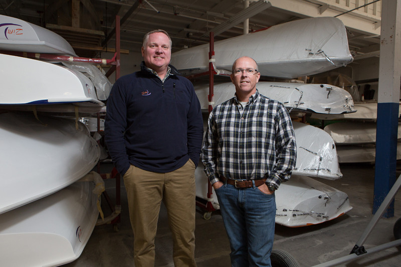 Two men stand smiling in a warehouse filled with several stacked and wrapped boat hulls. One man wears a navy pullover and khakis; the other wears glasses, a plaid shirt, and jeans. The background shows a well-lit, organized space.