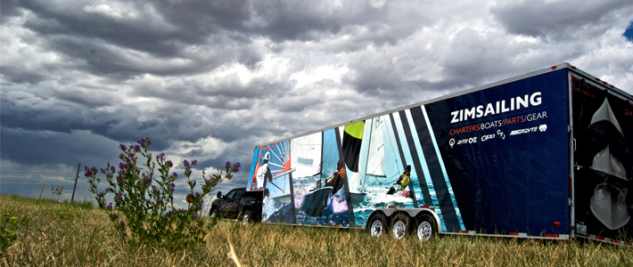 A large trailer labeled "Zim Sailing" sits in a grassy field under a dramatic, cloudy sky. The trailer features vibrant images of people sailing and lists boats and gear brands. Purple wildflowers are in the foreground.
