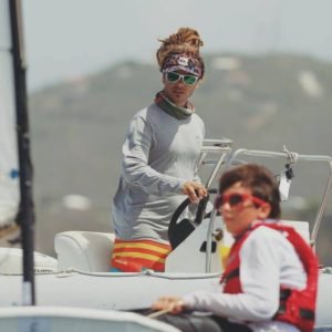 A person wearing sunglasses, a headband, and a long-sleeve shirt stands at the helm of a boat, while a child in a life jacket sits in the foreground. The background shows a blurred, hilly landscape.