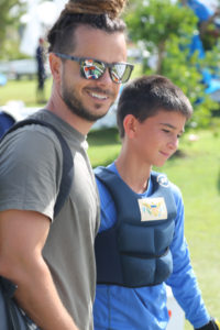 A man wearing sunglasses and a t-shirt stands smiling next to a boy in a blue long-sleeve shirt and padded vest outdoors on a sunny day. Trees and blurred people are visible in the background.