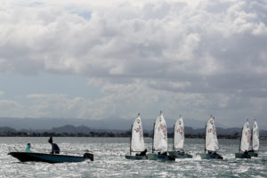 Several small sailboats with young sailors compete on the water, while a motorboat with two people follows nearby. The scene is set against a cloudy sky and distant mountains.