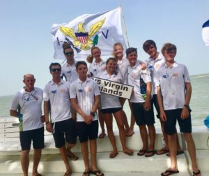 A group of ten people in matching white shirts stand outdoors by the water, holding a large U.S. Virgin Islands flag and a “US Virgin Islands” sign, smiling at the camera on a sunny day.