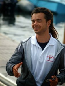 A smiling man with dreadlocks, wearing a gray zip-up hoodie over a white polo shirt, stands outdoors near water, extending his right hand as if to shake hands.