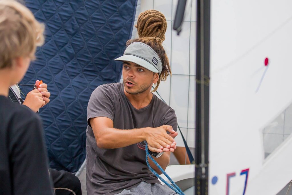 A person with dreadlocks and a visor cap gestures with their hands while talking to someone off-camera. They are sitting indoors near sailing equipment, appearing to explain or demonstrate something.