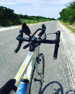 A close-up view of a white road bike’s handlebars on an empty, sunlit road lined with greenery on both sides, stretching into the distance under a blue sky.