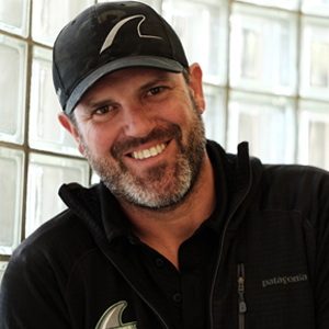 A man with a beard and mustache, wearing a black cap and black Patagonia jacket, smiles in front of a wall made of glass blocks.
