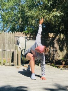 A person wearing a gray outfit and pink headband performs an outdoor stretch on a wooden deck, reaching one arm upward and the other arm touching the ground, with trees and a wooden fence in the background.