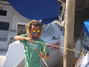 A person with dreadlocks, wearing sunglasses, a colorful face covering, and a green t-shirt, pulls a yellow rope on a docked boat with a blue cover in the background.