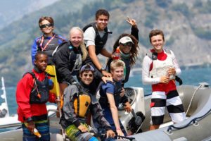 A group of smiling teenagers wearing life jackets pose together on an inflatable boat on a sunny day, with water and mountains visible in the background.