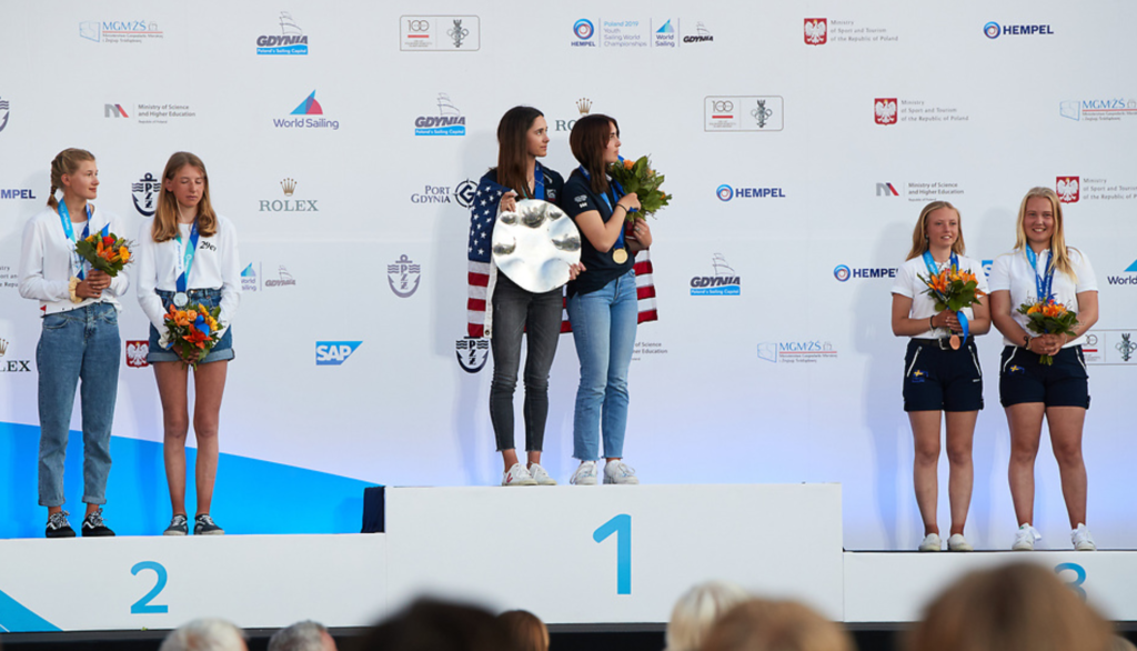 Three teams of young female athletes stand on a podium, holding medals and flowers. The winning team in the center holds a trophy, all smiling. The backdrop displays event and sponsor logos.