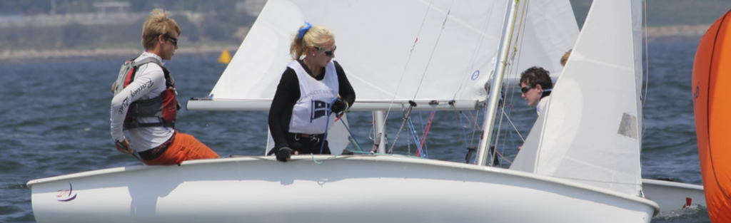 Two people wearing life jackets are sailing a small white sailboat on a calm body of water, with another sailboat and orange buoy visible in the background.