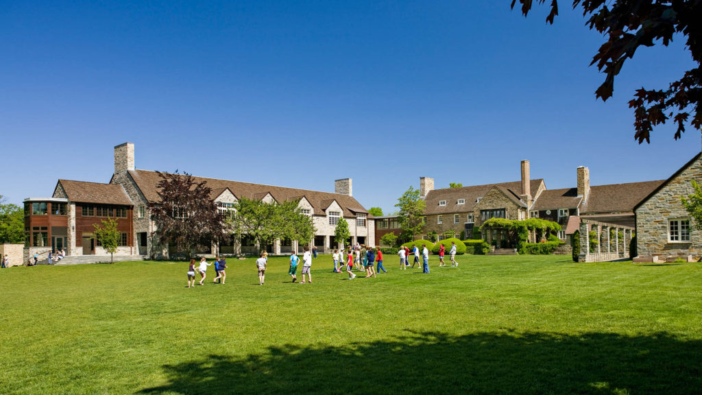 Students walk and play on a large grassy lawn in front of a stone and brick school building under a clear blue sky, with trees and landscaping around the campus.