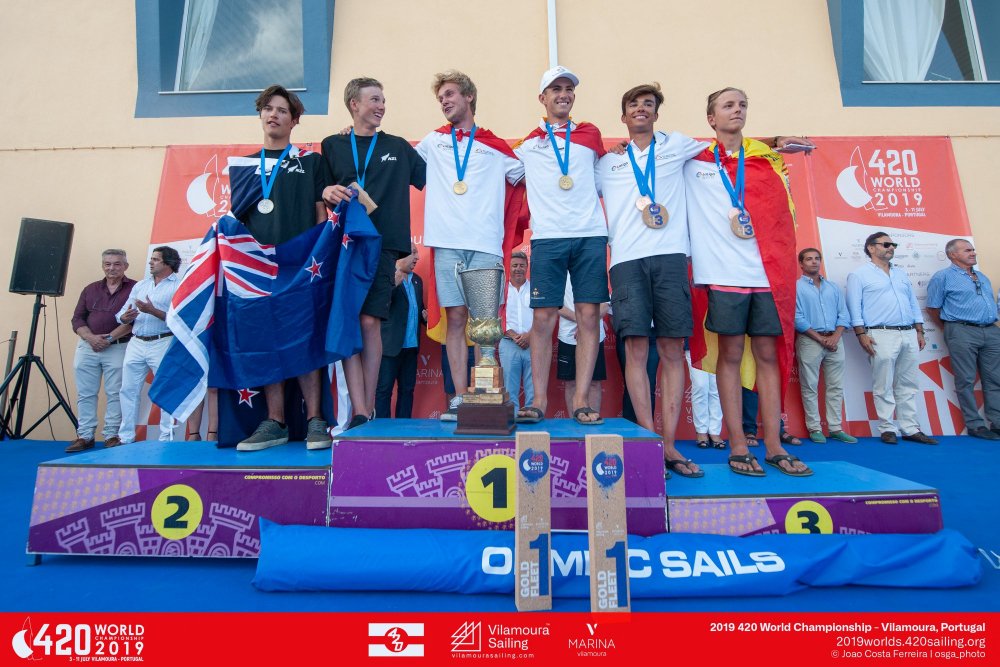 Six young male sailors stand on a podium with medals, trophies, and flags at the 420 World Sailing Championship 2019 awards ceremony in Vilamoura, Portugal. Smiling, they celebrate their achievements.