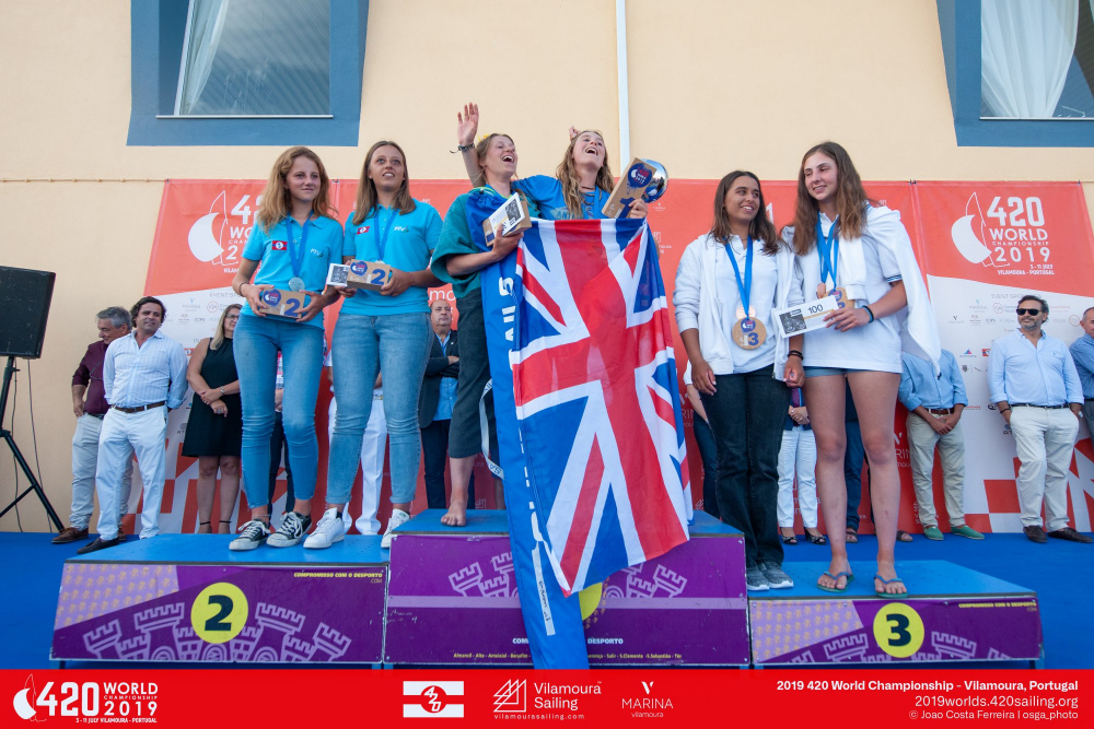 A group of female sailors stand on a podium at the 420 World Championship 2019 award ceremony. The winning team holds a British flag and gold medals, while runners-up display silver and bronze medals.