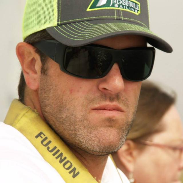 A man with stubble wears a neon green and black cap, dark sunglasses, and a yellow strap labeled "FUJINON." He looks serious, with a blurred person in the background.