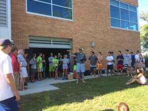 A group of people, mostly young adults, gather outside a brick building. One person stands in front, speaking and gesturing, while others stand or sit on the grass, listening attentively. It’s a sunny day.