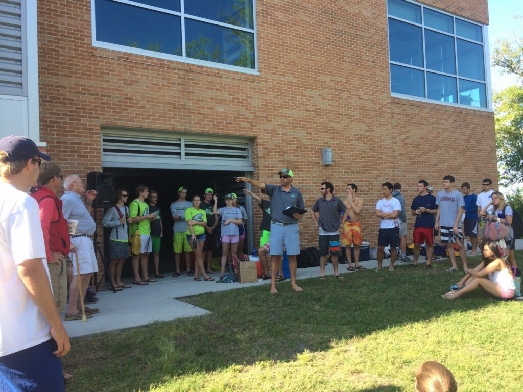 A group of people, mostly young adults, gather outside a brick building. One person stands in front, speaking and gesturing, while others stand or sit on the grass, listening attentively. It’s a sunny day.