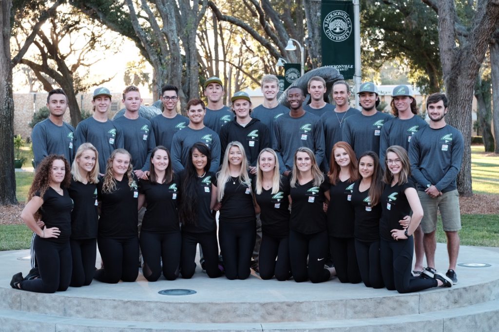 A group of young adults, some kneeling and some standing, pose outdoors in two rows, wearing matching black or gray shirts with a green logo, in front of trees and a university banner.