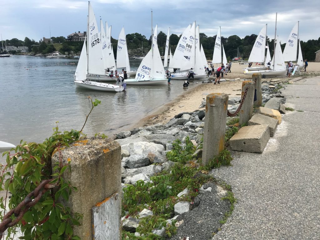 Several white sailboats with visible numbers are docked at a sandy beach, with people preparing them. Rocky shoreline, greenery, and a cloudy sky are in the background; a concrete walkway is in the foreground.
