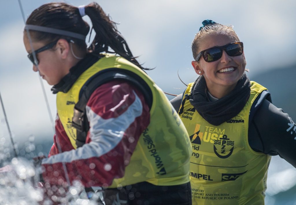 Two women wearing sunglasses and yellow vests with sailing logos are on a boat. One is smiling brightly at the camera, while the other looks focused on her task, with water and sky visible in the background.