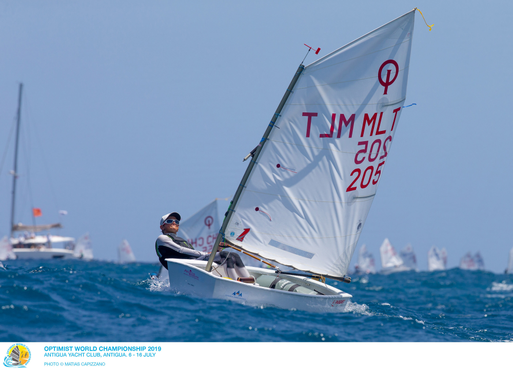 A young sailor competes in a small sailboat during the Optimist World Championship 2019, navigating choppy blue waters with other sailboats and a yacht visible in the background.