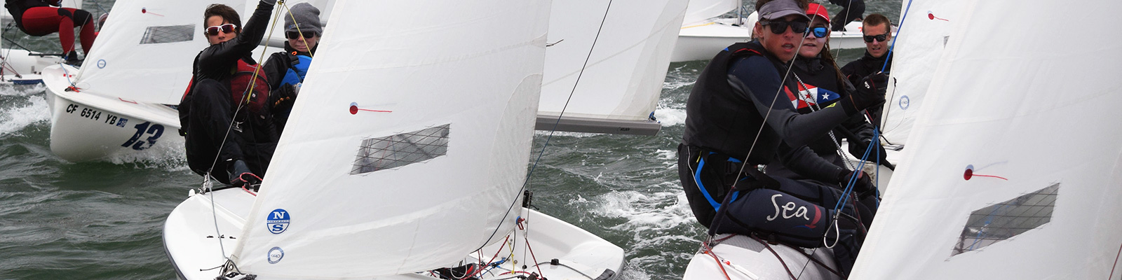 Several people wearing life jackets and sunglasses are sailing small boats in choppy water, closely maneuvering around each other during a sailing competition or regatta.