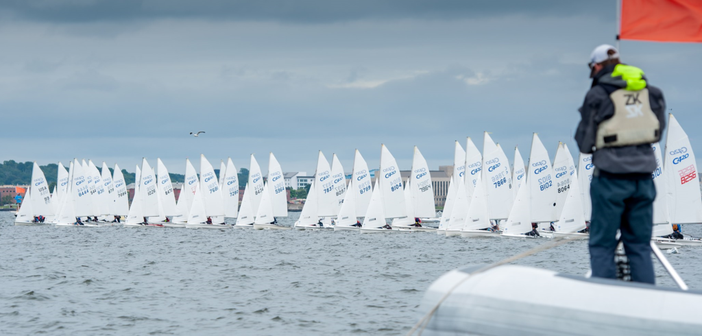A fleet of sailboats with white sails lined up on the water at the start of a race, with a person in a life vest standing on an inflatable boat in the foreground, holding a red flag.