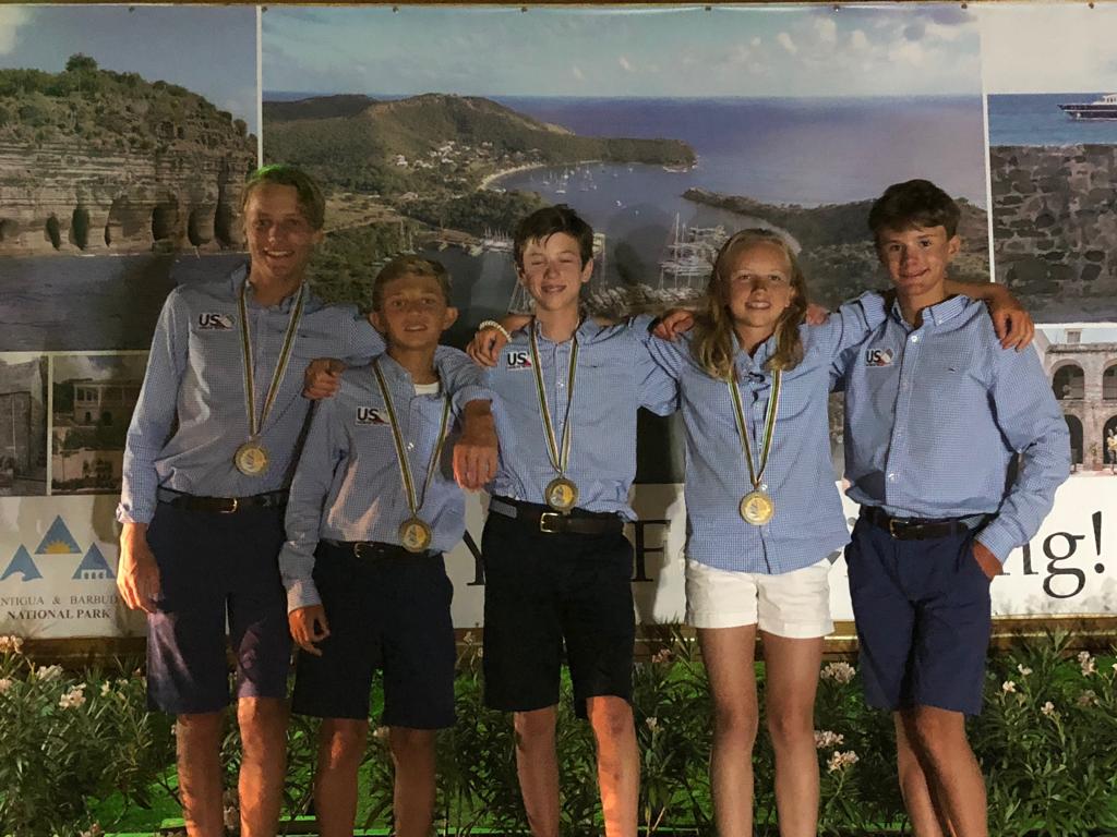 Five young people wearing blue shirts, navy shorts, and gold medals stand smiling in front of a scenic backdrop with coastal landscapes. They have their arms around each other and appear happy and celebratory.