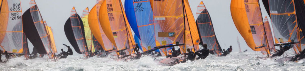 A group of sailboats with vibrant, colorful sails race closely together on choppy water, with sailors leaning out over the edge as waves splash around them.