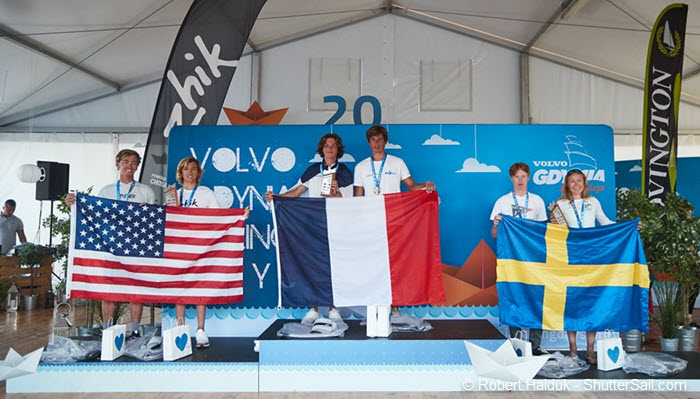 Five young sailors stand on a podium holding the flags of the USA, France, and Sweden at an awards ceremony, with event banners and medals visible in the background.