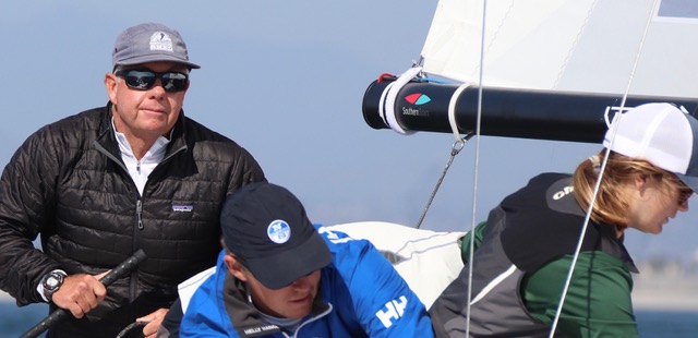 Three sailors work together on a sailboat; one man in a black jacket and sunglasses steers, while two others in blue and black jackets handle ropes and equipment near the sail.