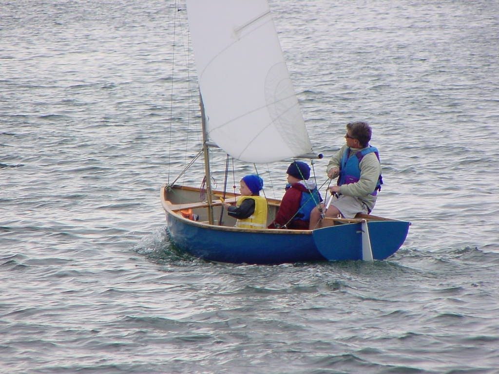 Three people, two children and an adult, are sailing a small blue sailboat on a body of water. They are all wearing life jackets, and the adult is steering the boat. The sail is raised and the water is calm.