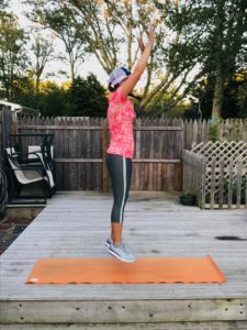 A person in athletic clothes and a cap is outdoors, mid-jump on an orange exercise mat on a wooden deck, with arms raised overhead. Trees and a wooden fence are visible in the background.