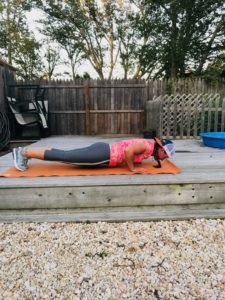 A woman wearing a pink tank top, gray leggings, and a cap does a plank exercise on an orange mat outdoors on a wooden deck, with a fenced yard and trees in the background.