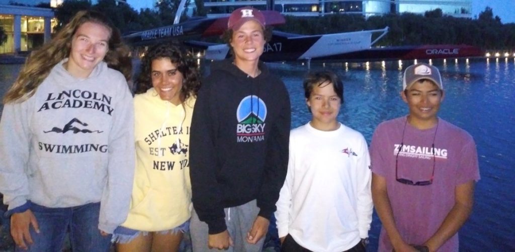 Five smiling teens in casual hoodies and T-shirts stand together by the water at dusk, with a large racing sailboat and buildings visible in the background.