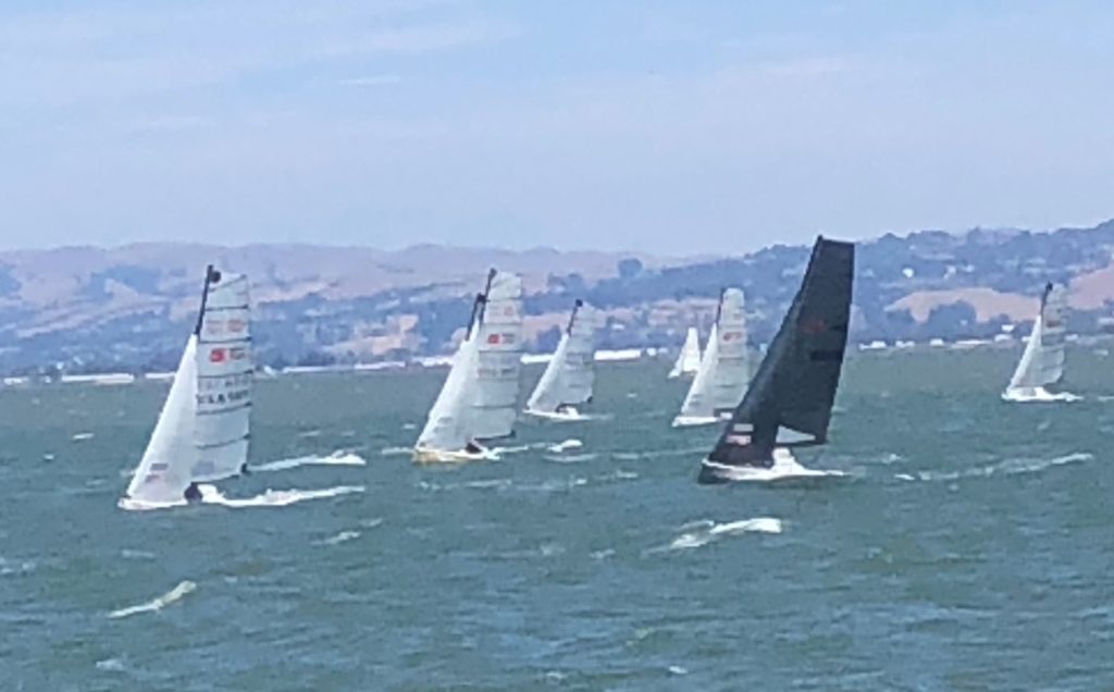 Several sailboats racing on choppy water with hilly land and houses in the background; most sails are white, but one is black. The sky is partly cloudy.
