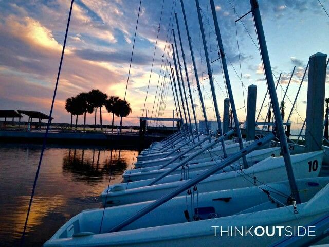 A row of sailboats lined up by a dock at sunset, with palm trees silhouetted in the background and colorful clouds reflecting on the calm water. The words 'THINK OUTSIDE' appear in the bottom right corner.