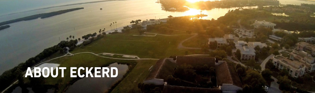 Aerial view of a scenic waterfront campus at sunset with green fields, buildings, and water; text "ABOUT ECKERD" appears in bold white letters on the lower left.