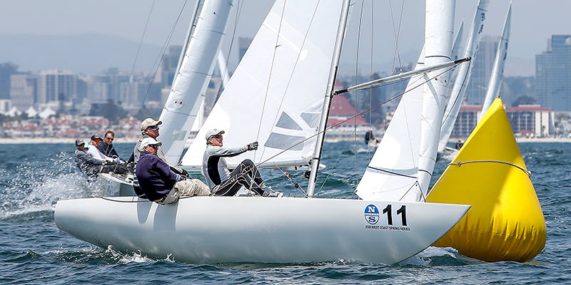 Four sailors race a white sailboat marked with the number 11 around a bright yellow buoy on the water, with city buildings and a coastline visible in the background.