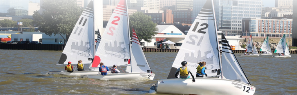 Sailboats with numbered sails compete on a river, with several people on each boat. City buildings and trees are visible in the background under a bright, clear sky.