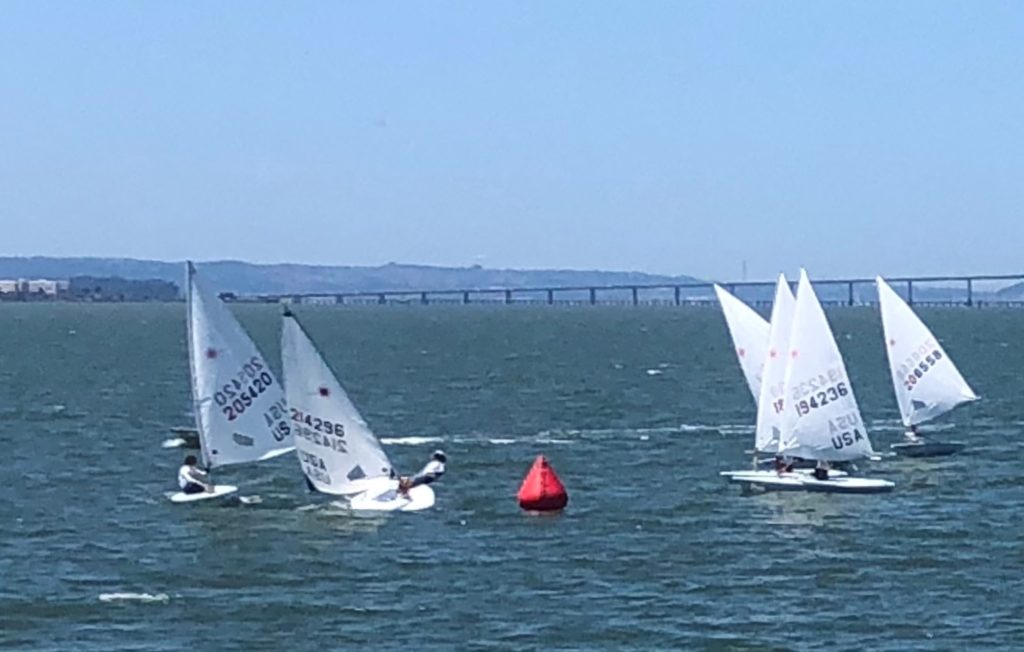 Four small sailboats race around a red buoy on a sunny day, with choppy blue water and a distant bridge visible in the background. Sailors are adjusting their sails as they navigate the course.