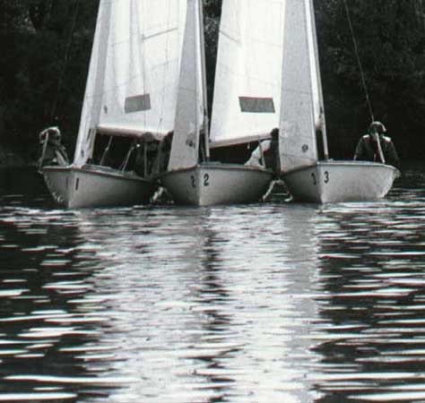Three small sailboats with the numbers 1, 2, and 3 on their bows are close together on calm water, with several people visible on each boat and trees in the background.