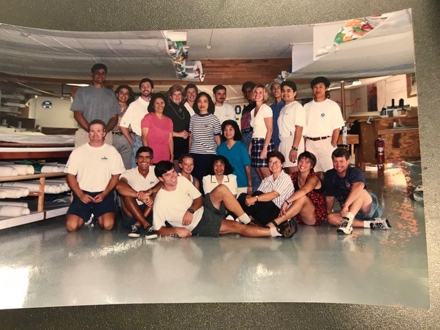 A group of about 20 people, casually dressed, pose together and smile inside what appears to be a workspace or workshop with tables, shelves, and supplies in the background.