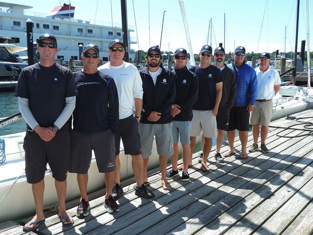 Nine men stand in a row on a wooden dock in front of sailboats, wearing casual sailing attire including sunglasses, shorts, caps, and jackets, with a marina and large boat in the background on a sunny day.
