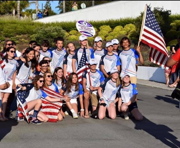 A group of young people wearing matching USA shirts and hats pose outdoors with several American flags, smiling and gathered closely together in the sunlight. Green bushes and a white wall are in the background.